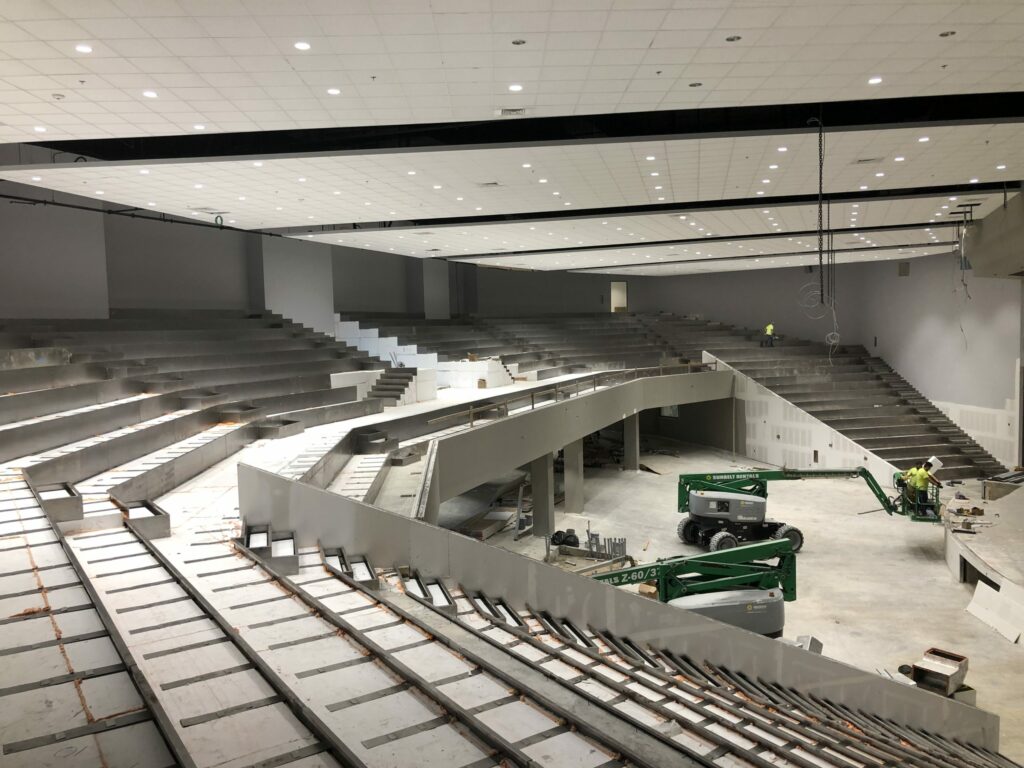 Large auditorium under construction with tiered seating, ceiling lighting installed, and workers using green boom lifts for finishing work.