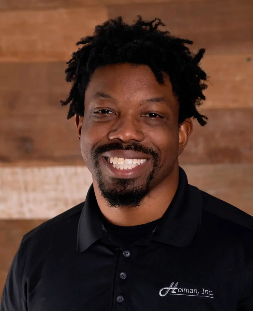 Devon, team member at Holman, Inc., wearing a black company polo shirt and standing in front of a wood panel wall.