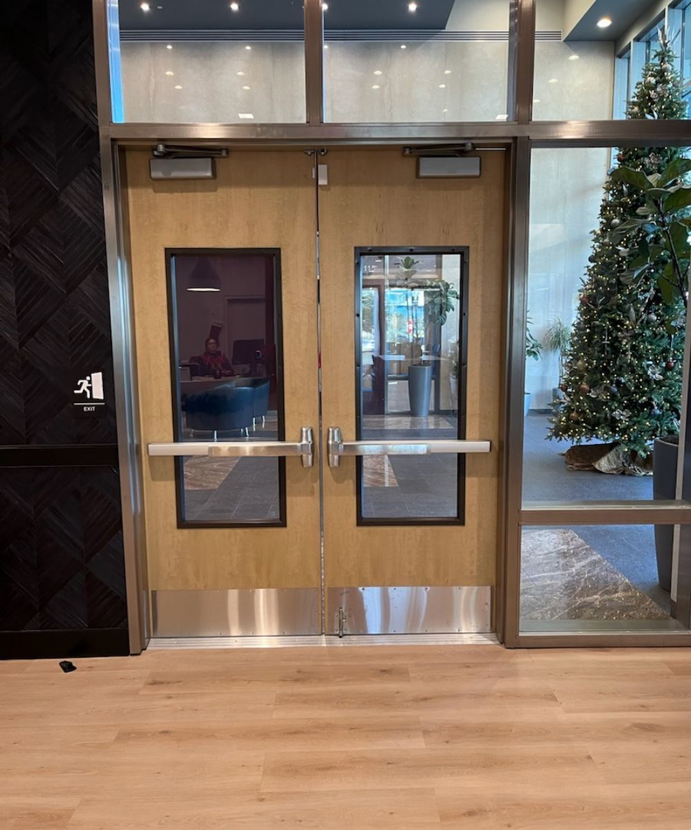 Double wooden doors with glass panes and metal push bars in a lobby, holiday tree visible outside. Start of an exit doorway in a modern building.