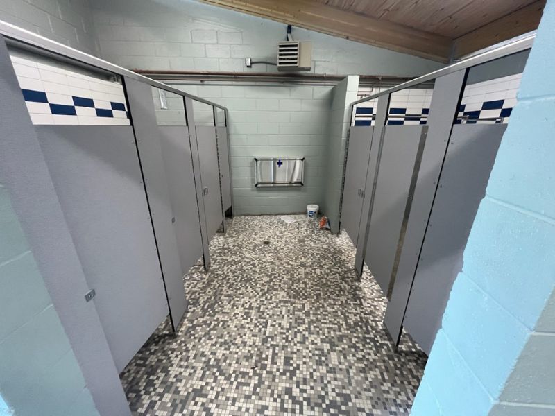phenolic-toilet-partitions-locker-room-restroom-jacksonville-fl - Holman, Inc. Public locker room with rows of gray stall doors, blue-white tile border, and a mosaic floor; back wall has a wall-mounted dispenser and a few cleaning items on the floor.