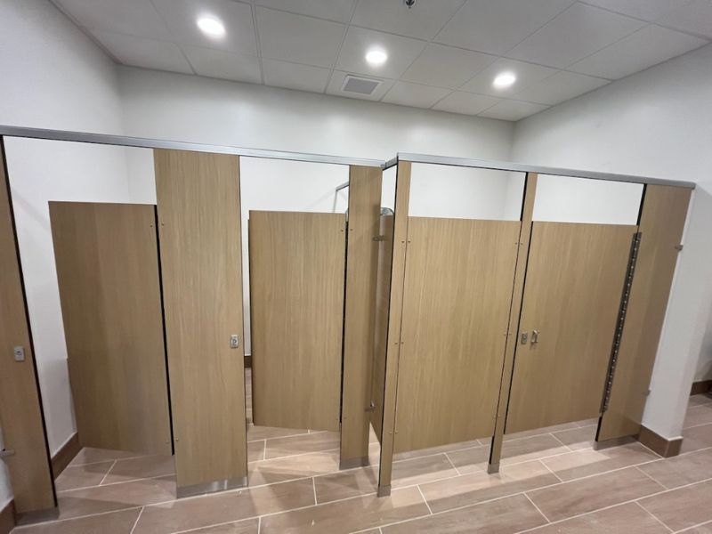 wood-look-compact-laminate-toilet-partitions-jacksonville-fl - Holman, Inc. Public restroom with row of wooden stall doors and tiled floor, some doors slightly ajar, bright ceiling lights above.