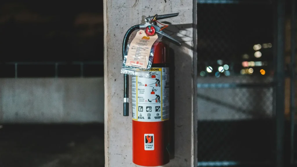 Red fire extinguisher mounted to a concrete pillar with instruction tag in a dim indoor setting